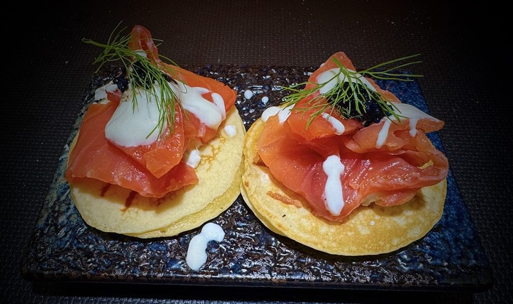Blinis con salmone marinato, panna acida e caviale, serviti su una piastra di ceramica scura